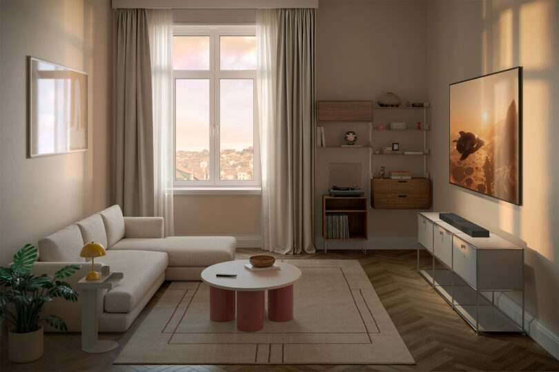 A modern living room with a white sofa, round coffee table, TV on the wall, KEF XIO speakers on shelves, a window with city view, and neutral-colored decor.