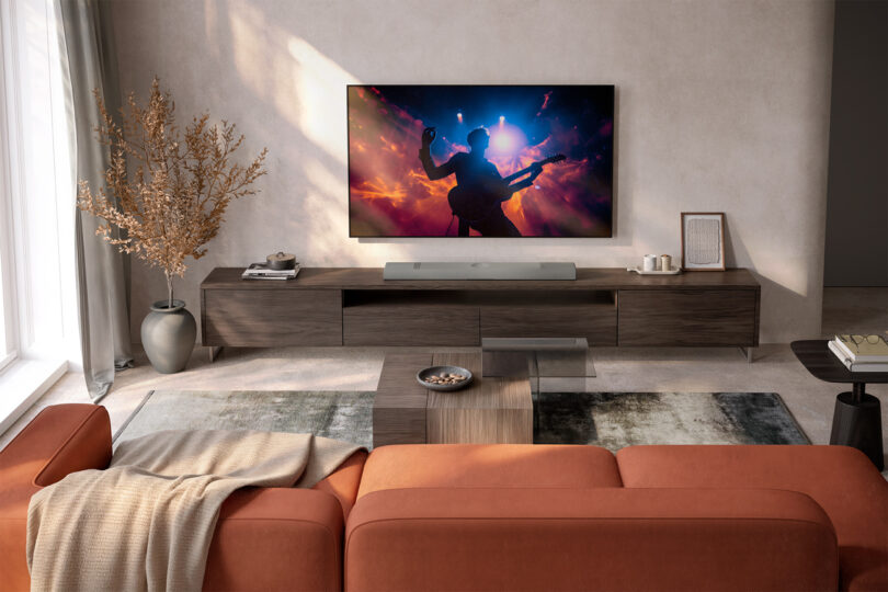 A modern living room with an orange sofa, wooden furniture, a KEF XIO speaker beside the large TV displaying a concert scene, and a potted plant by the window.