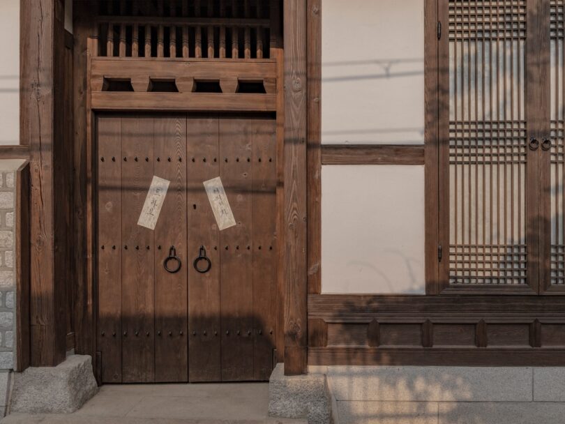 Traditional wooden double doors with metal rings and paper notices, beside a wooden lattice window, on a building with light-colored walls and stone steps.
