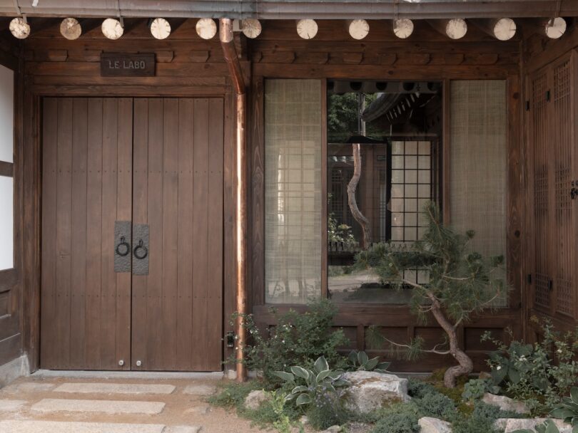 Wooden double doors labeled "Le Labo" beside a large window with bamboo blinds, small green plants, and stone landscaping in front of a traditional-style building.