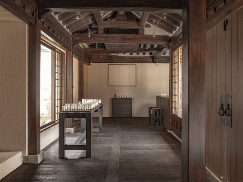 Una habitación minimalista con pisos y vigas de madera, ventanas grandes y una mesa con cristalería, con paredes neutrales y muebles simples.