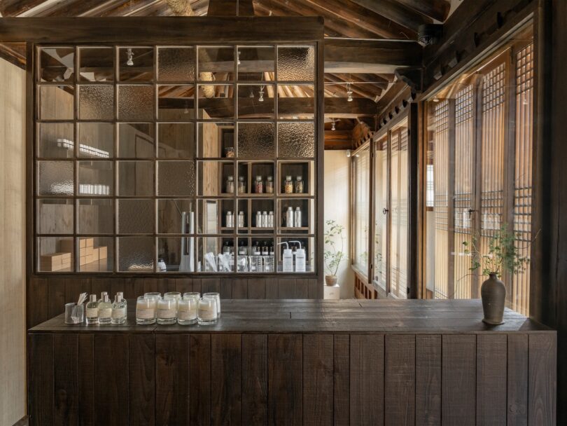 A minimalist shop interior with wooden beams, a dark wood counter displaying candles and bottles, glass shelving, and sunlight streaming through wooden lattice windows.