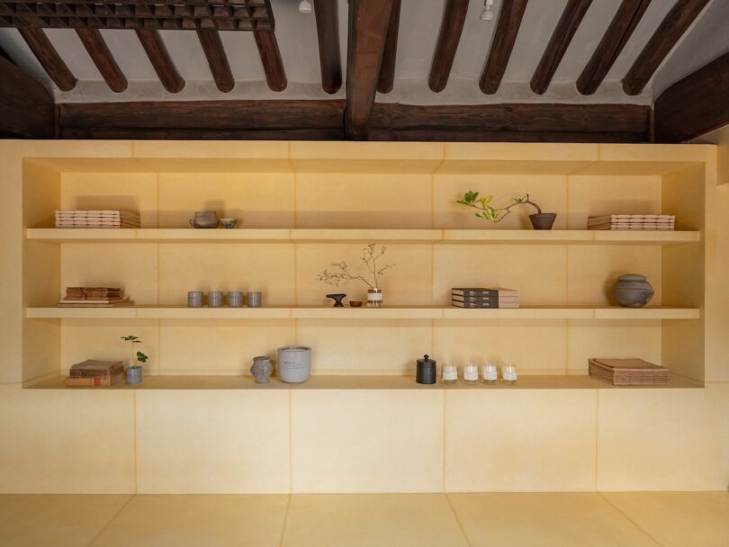 Minimalist shelving with decorative objects, books, ceramics, plants, and neutral tones, set against a yellow wall with exposed wooden ceiling beams above.