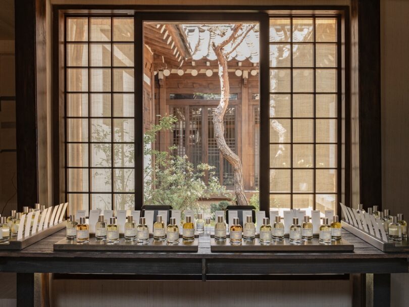 Glass bottles of perfume are arranged on a wooden table in front of a large window with a view of a traditional wooden courtyard and tree outside.
