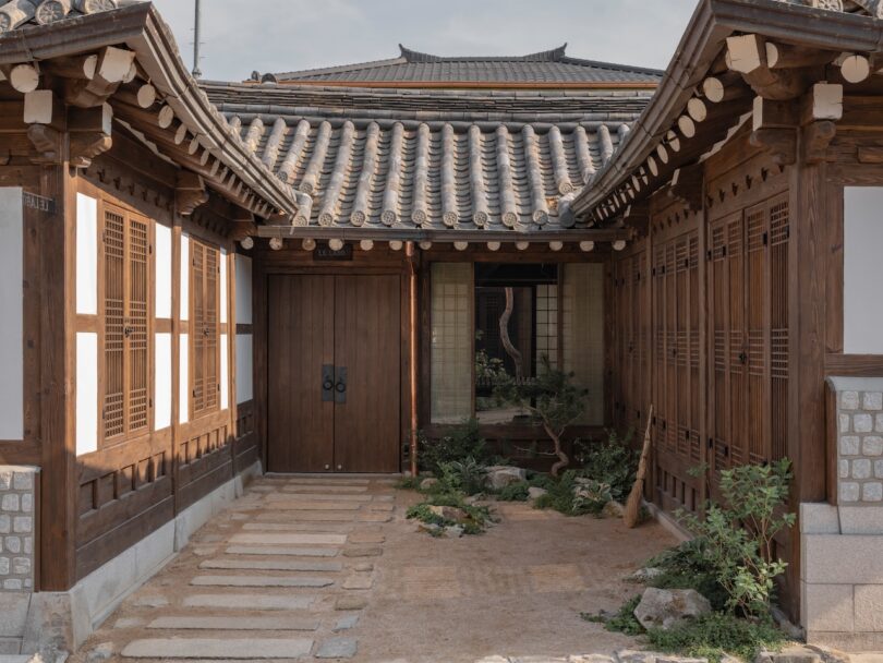 Traditional Korean hanok house courtyard with wooden doors, tiled roof, stone pathway, and small plants along the edges.