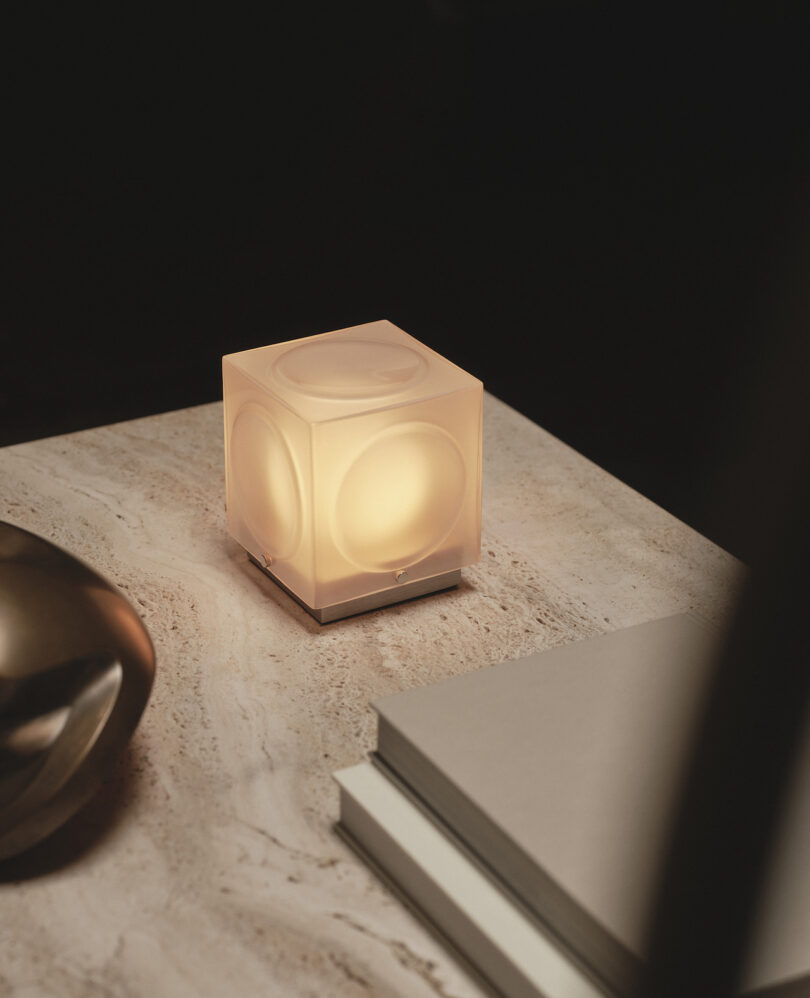 A small cube-shaped lamp with a glowing light sits on a marble surface next to stacked books and a metallic decorative object