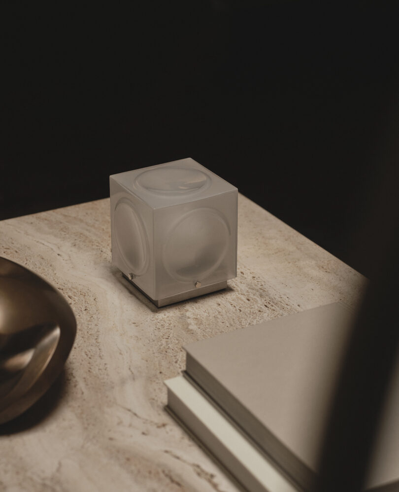 A small cube-shaped lamp sits on a marble surface next to stacked books and a metallic decorative object