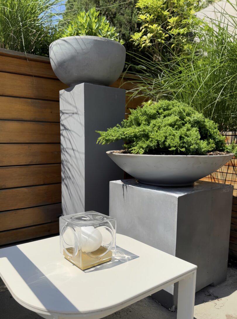 A modern outdoor setting with two concrete planters on gray pedestals containing green plants, next to a white table holding a glass decorative object