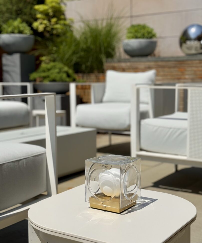 Modern outdoor patio with white cushioned chairs, a white table, and a decorative glass cube centerpiece; potted plants and a metallic sphere in the background