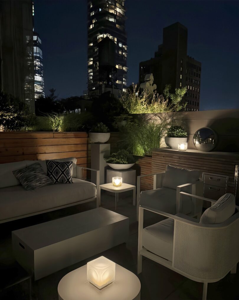 Rooftop patio at night with modern outdoor furniture, potted plants, and cube-shaped lanterns; city buildings are visible in the background