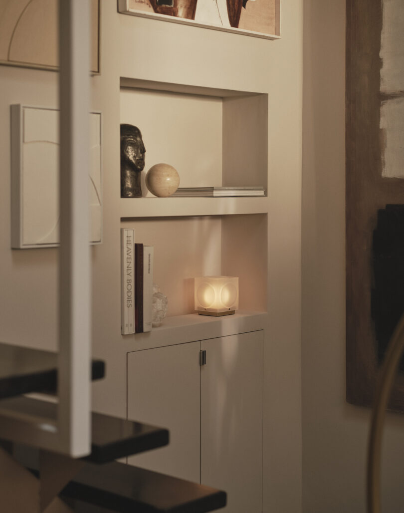 A built-in wall shelf displays books, art, a sculpture, and a glowing spherical lamp in a softly lit, modern interior