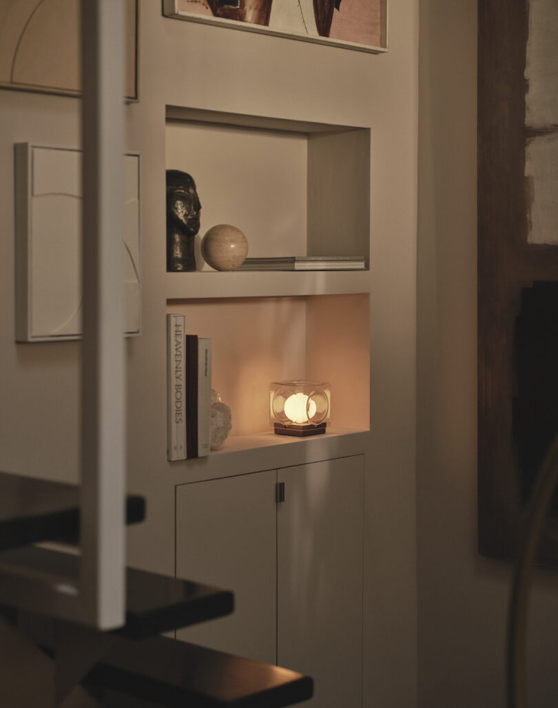 A built-in wall shelf displays books, art, a sculpture, and a glowing spherical lamp in a softly lit, modern interior