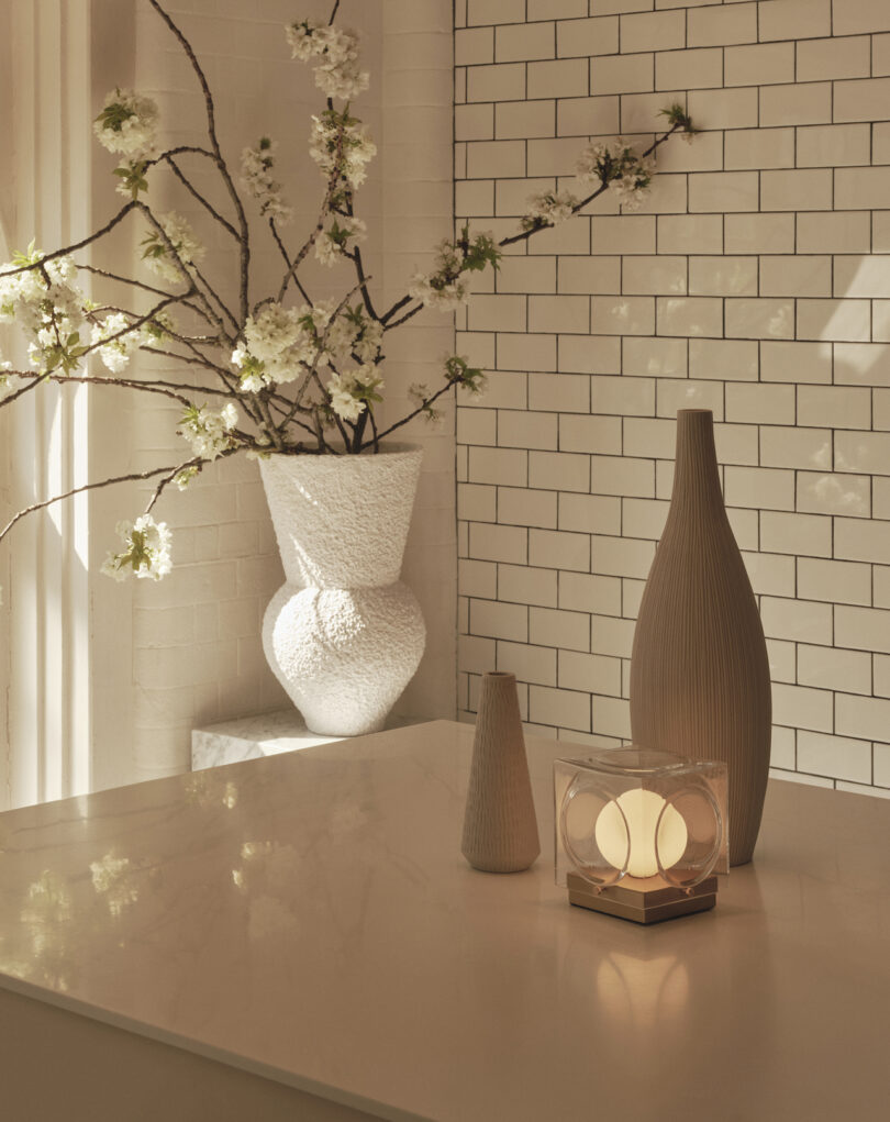 A white vase with flowering branches, two ribbed ceramic vases, and a cube-shaped lamp sit on a marble countertop against a white subway tile wall