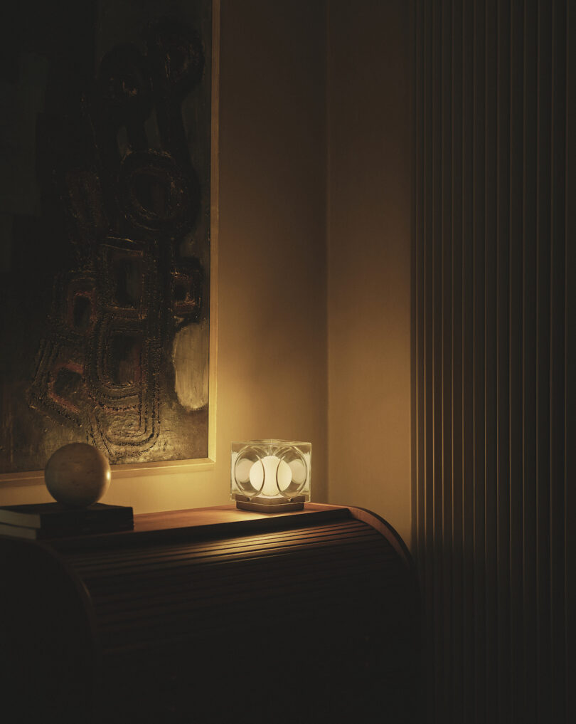 A cube-shaped lamp emits warm light on a wooden sideboard with books and a round object, next to a textured abstract painting and vertical radiator