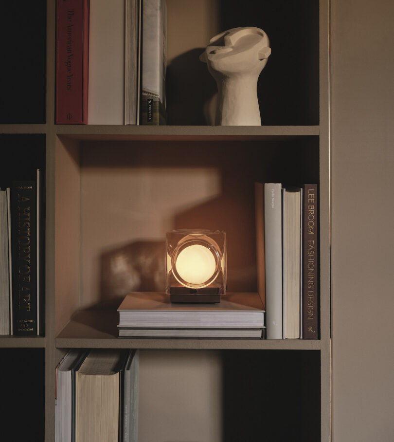 A bookshelf with neatly arranged books, a modern table lamp emitting warm light, and a white abstract sculpture on the upper shelf