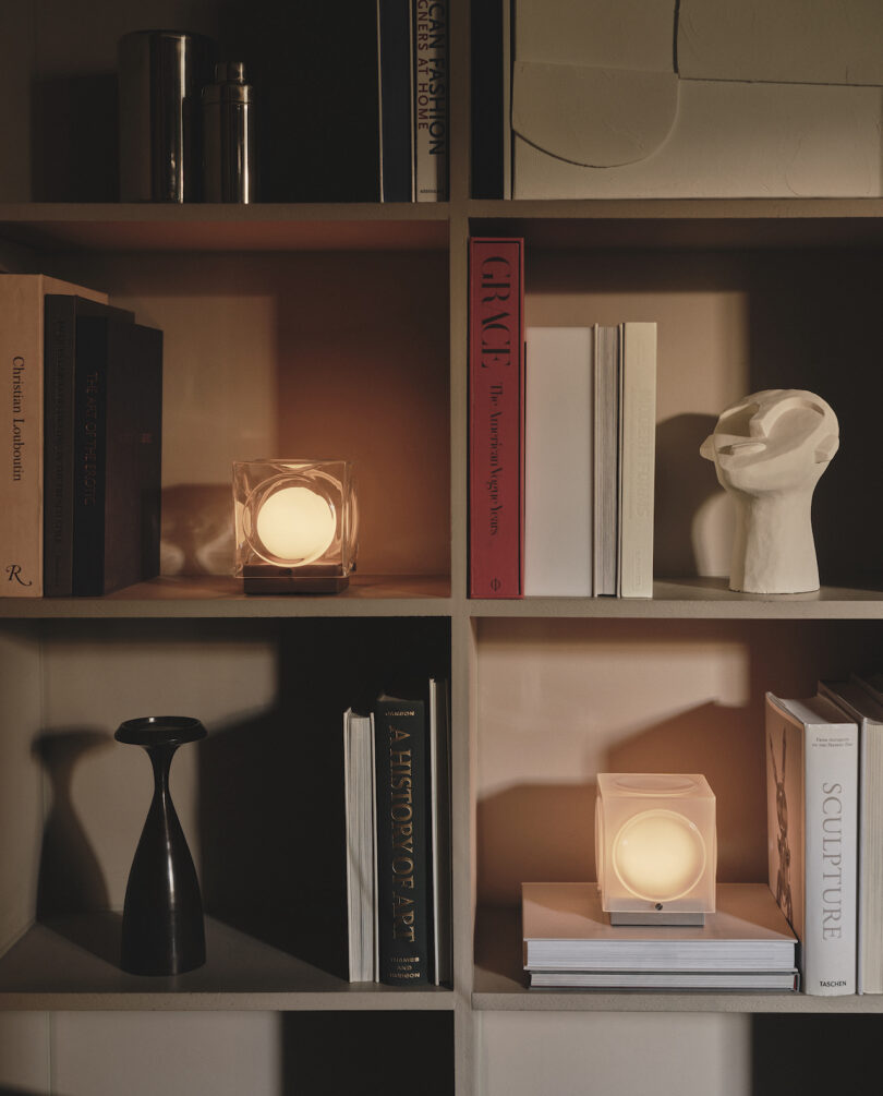 A bookshelf with art books, two glowing cube lamps, a black vase, and a white abstract sculpture head on display