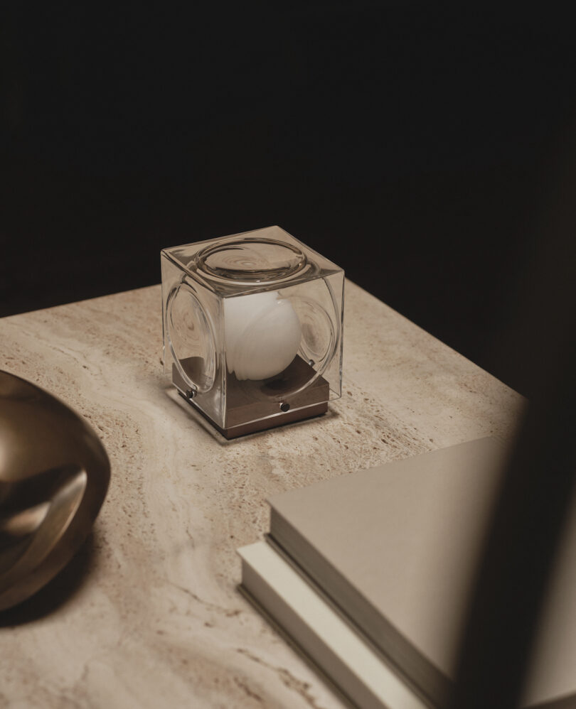 A small cube-shaped lamp sits on a marble surface next to stacked books and a metallic decorative object