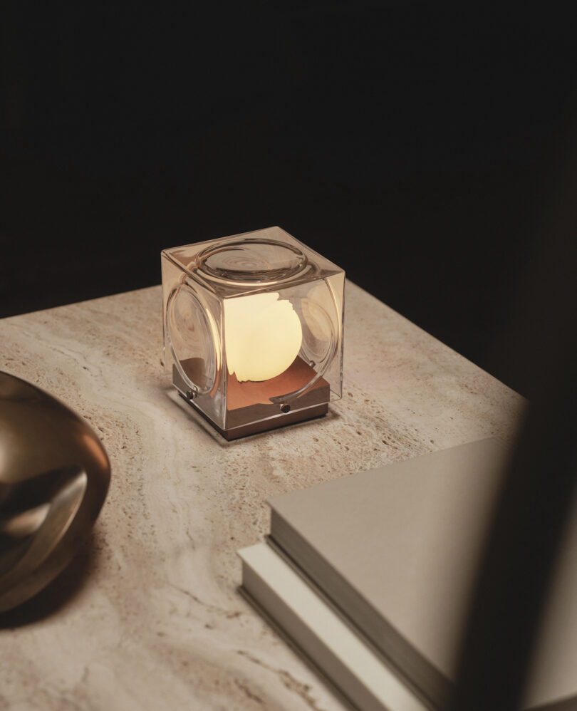 A small cube-shaped lamp with a glowing light sits on a marble surface next to stacked books and a metallic decorative object