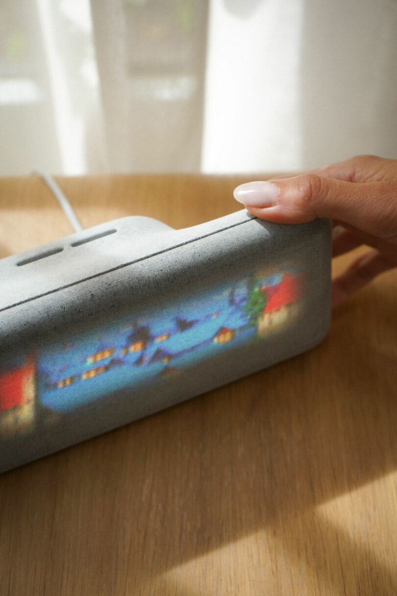 A hand adjusts a gray device showing a blurred colorful image on its screen, placed on a wooden surface near a window.