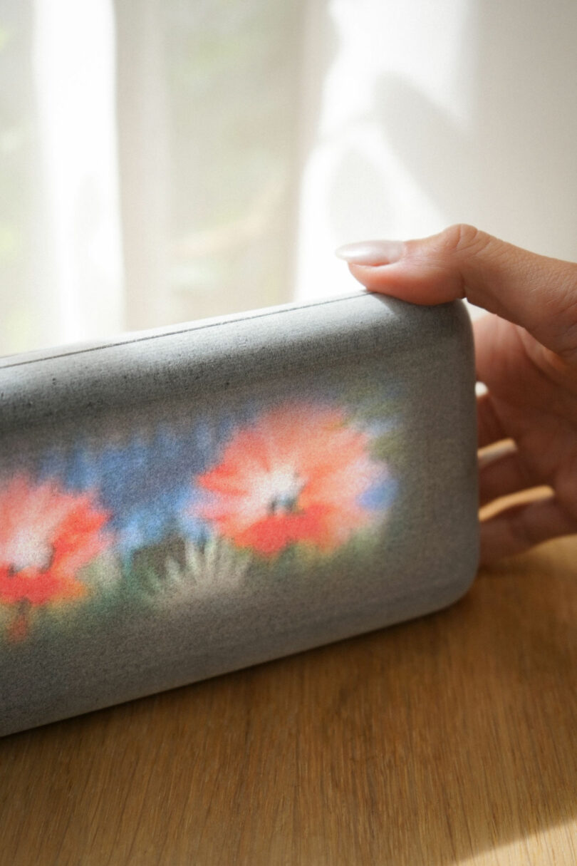 A hand holds a rectangular, gray devoce with a blurred floral design, resting on a wooden surface near a softly lit window.