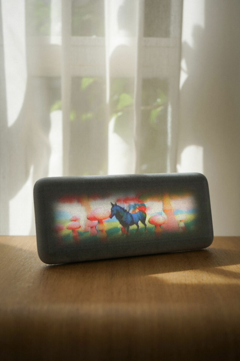 A rectangular device with a colorful image of a unicorn and mushrooms sits on a wooden surface in front of a sunlit window with sheer curtains.