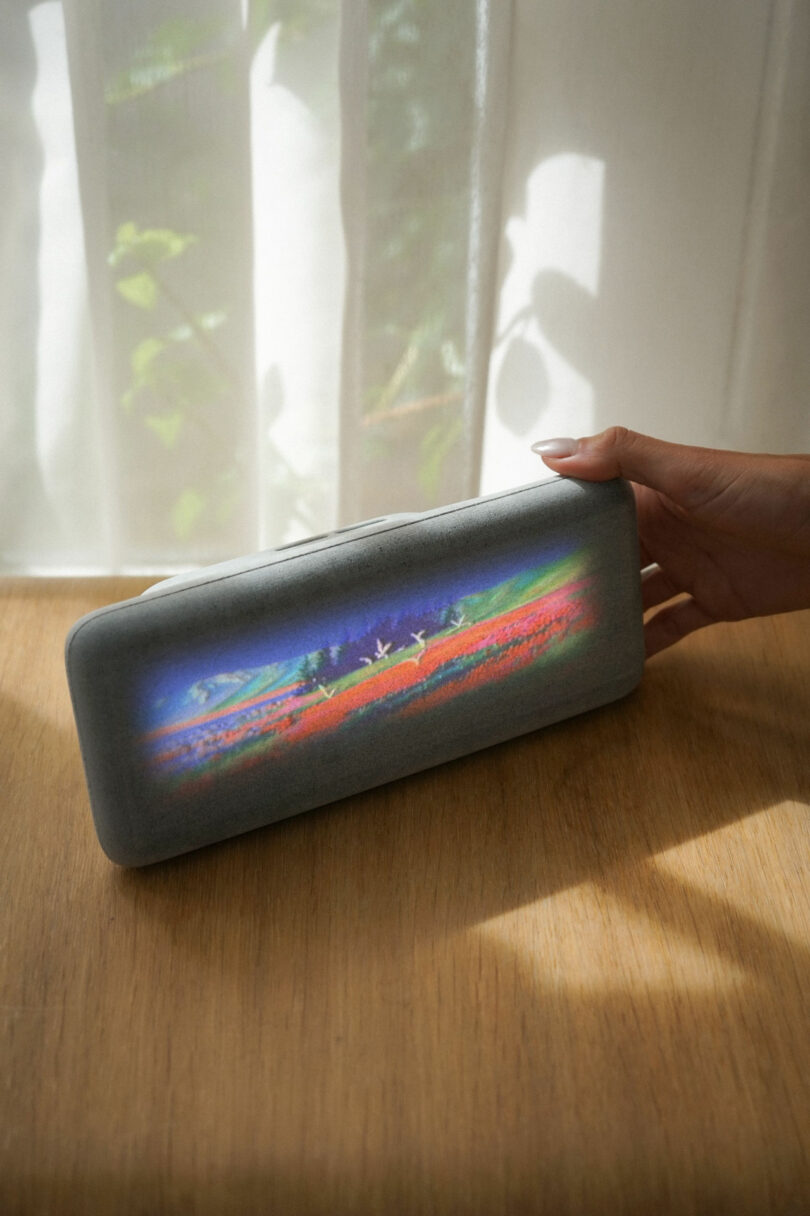 A hand holds a rectangular gray device with a colorful landscape image displayed on its front, placed on a wooden table near a sunlit window with sheer curtains