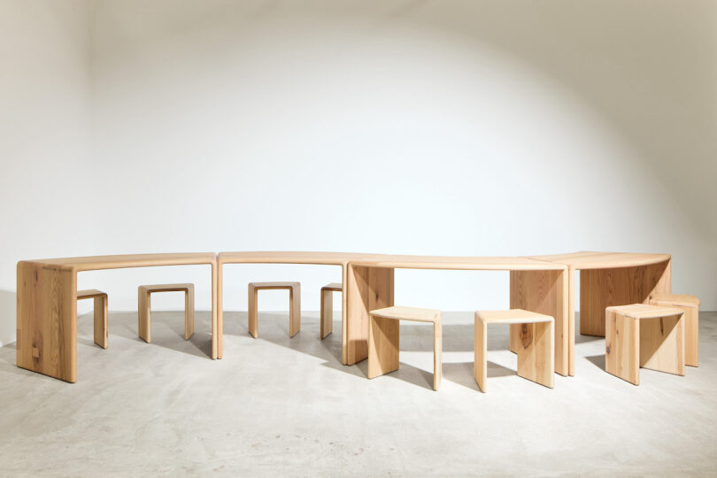 A minimalist wooden furniture set features several benches and stools with clean, angular lines, arranged in a curved formation in a bright, empty room
