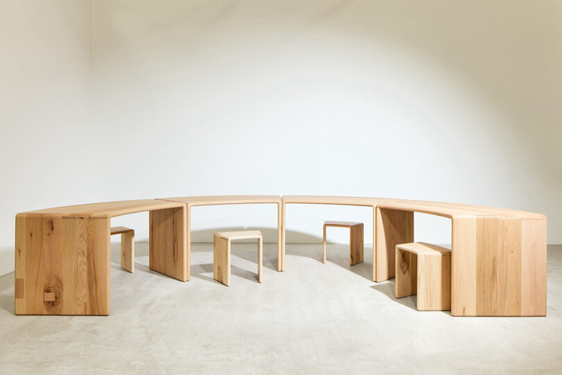 A set of minimalist wooden furniture with curved benches and small rectangular stools arranged in a semi-circle on a plain concrete floor