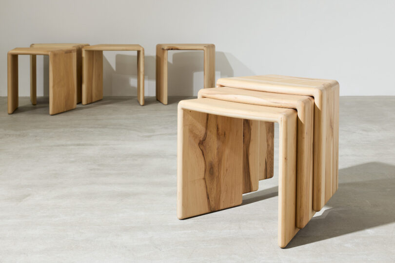 Three wooden nesting tables are stacked together in the foreground, with two more similar tables placed apart in the background on a concrete floor