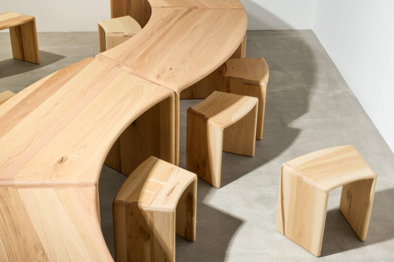 Curved wooden tables and matching wooden stools arranged on a light gray concrete floor against white walls