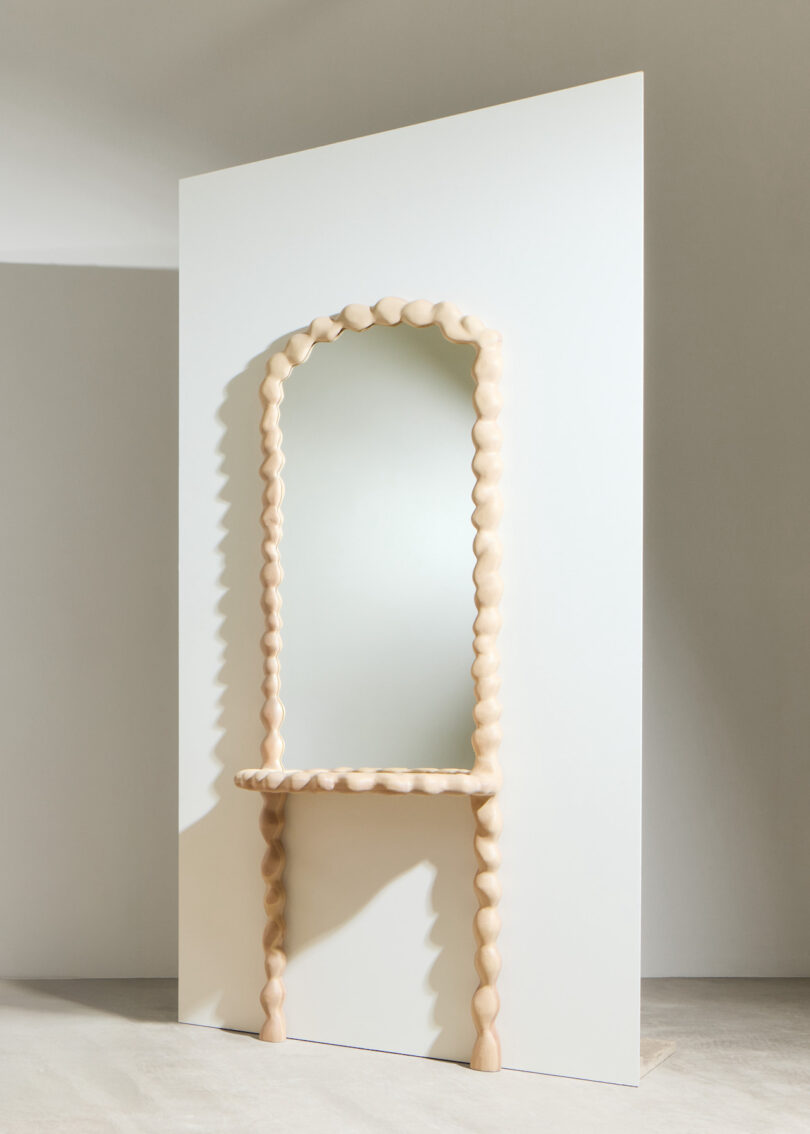 A tall rectangular mirror with a wavy, sculpted wooden frame and two matching wooden legs, positioned against a plain white background