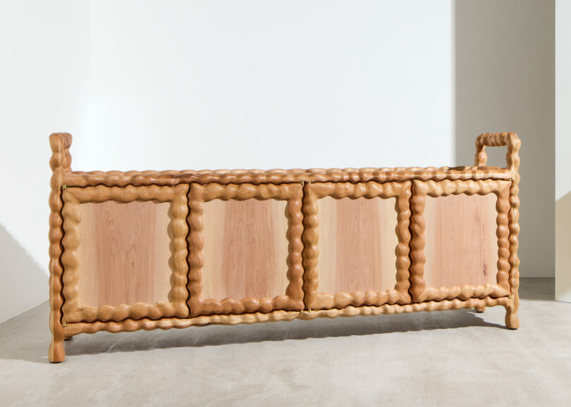 A wooden sideboard with four doors and a distinctive wavy, sculpted frame, set against a white wall on a concrete floor