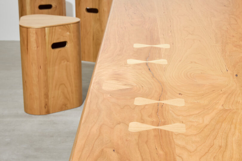 Close-up of a wooden table with butterfly joint inlays and two matching wooden stools with handholds, set on a light gray floor
