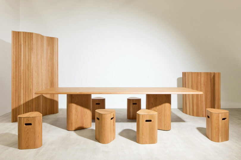 A minimalist wooden dining set with a rectangular table, eight round stools with handle cutouts, and two wood panel room dividers in a bright, neutral space