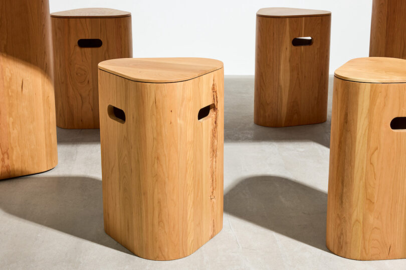 Several wooden stools with rounded triangular tops and side handle cutouts are arranged on a light gray floor in a well-lit room