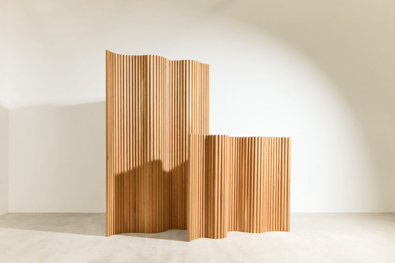 A wooden room divider with a wavy, ribbed design stands on a light-colored floor against a plain white wall