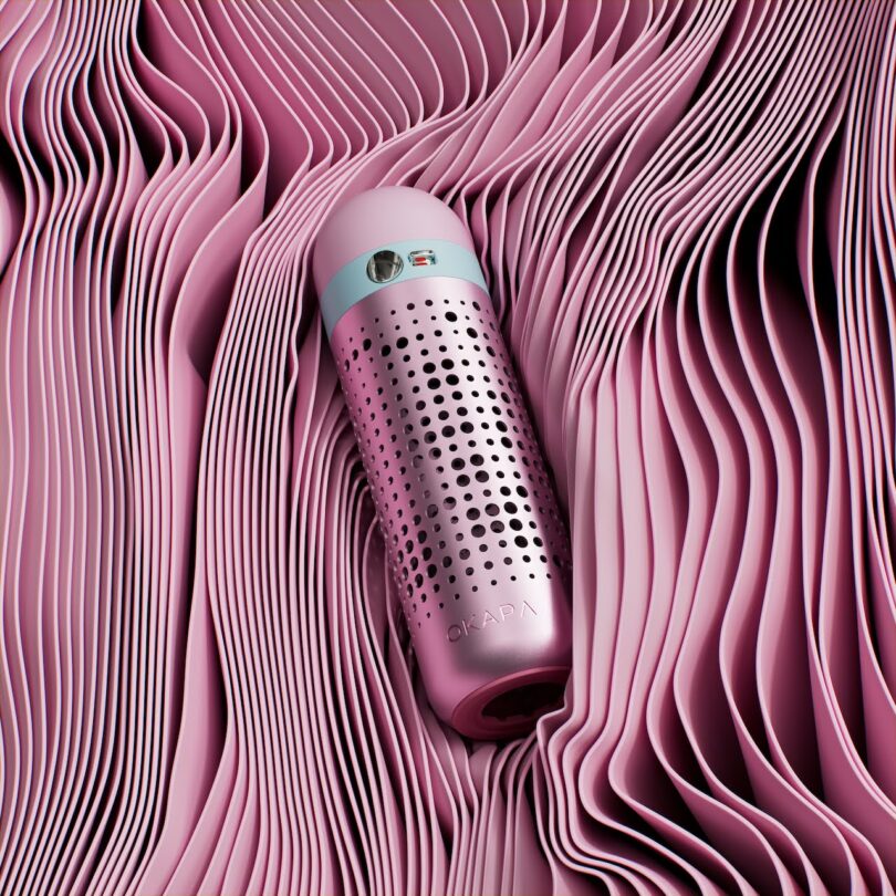 A pink metallic water bottle with vented holes and a blue band lies on wavy, layered sheets of pink material