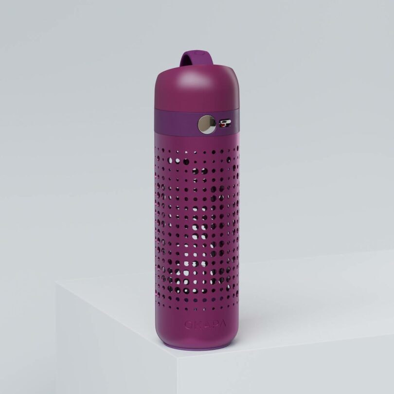 A purple water bottle with a perforated outer sleeve and a flip-top lid sits on a white surface against a light gray background