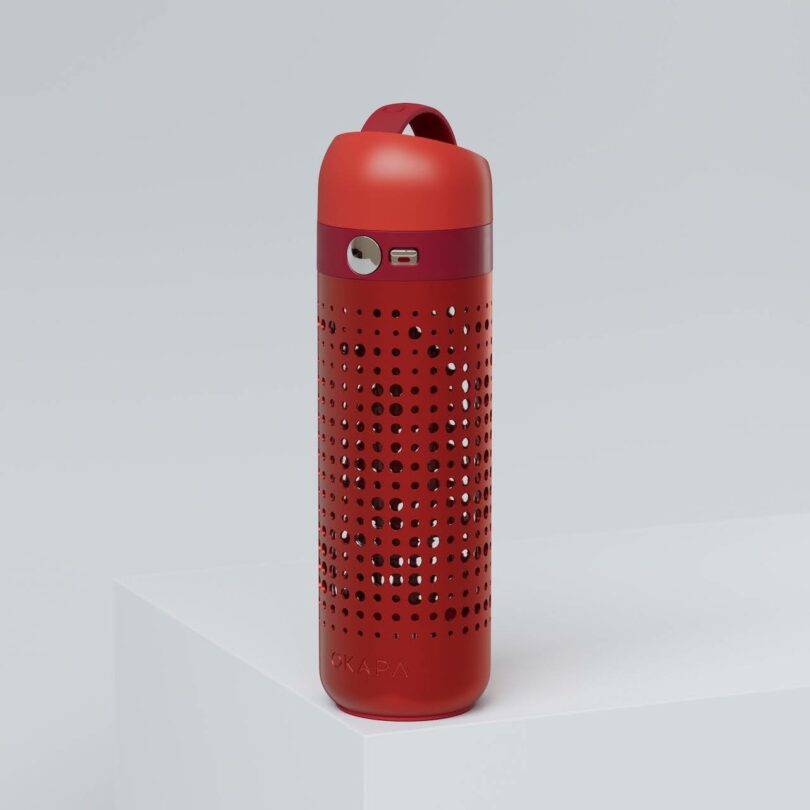 A red portable speaker with a perforated design and a carrying loop, placed on a white surface against a plain background