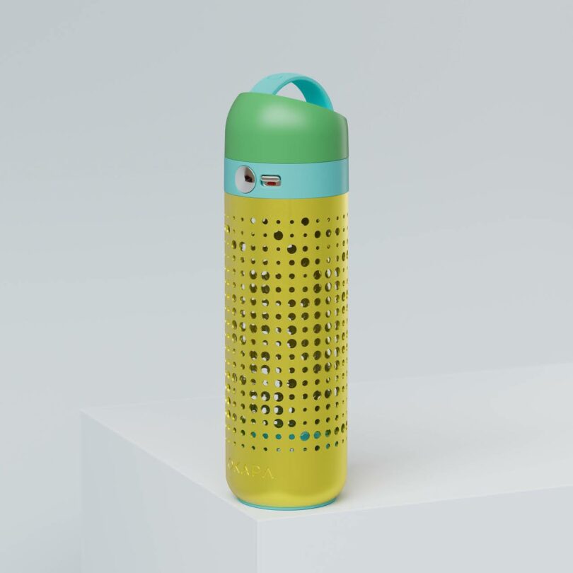 A green and yellow portable speaker with a blue accent, featuring a perforated design and visible charging port, placed on a white surface against a light background