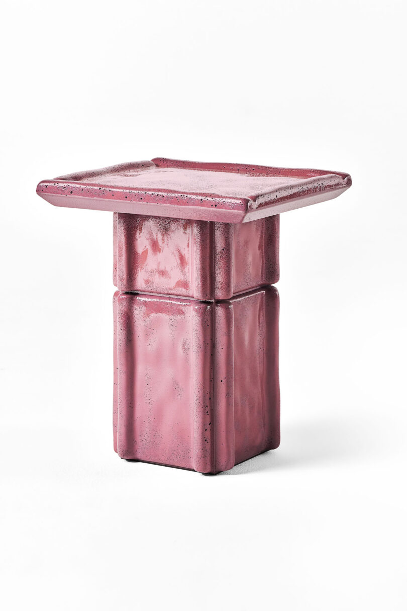 A small, square, pink table with a flat top and a blocky, textured pedestal base, set against a plain white background