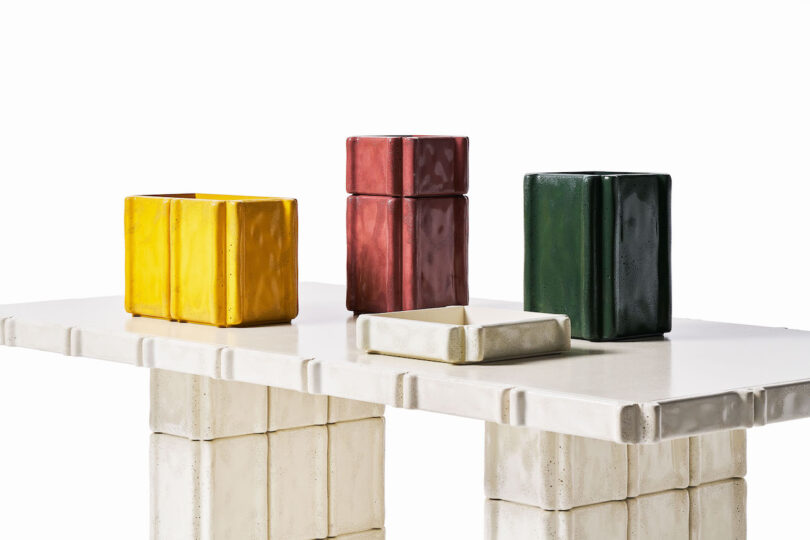 Four geometric ceramic containers in yellow, red, green, and cream sit on a matching cream-colored rectangular table with block-like legs