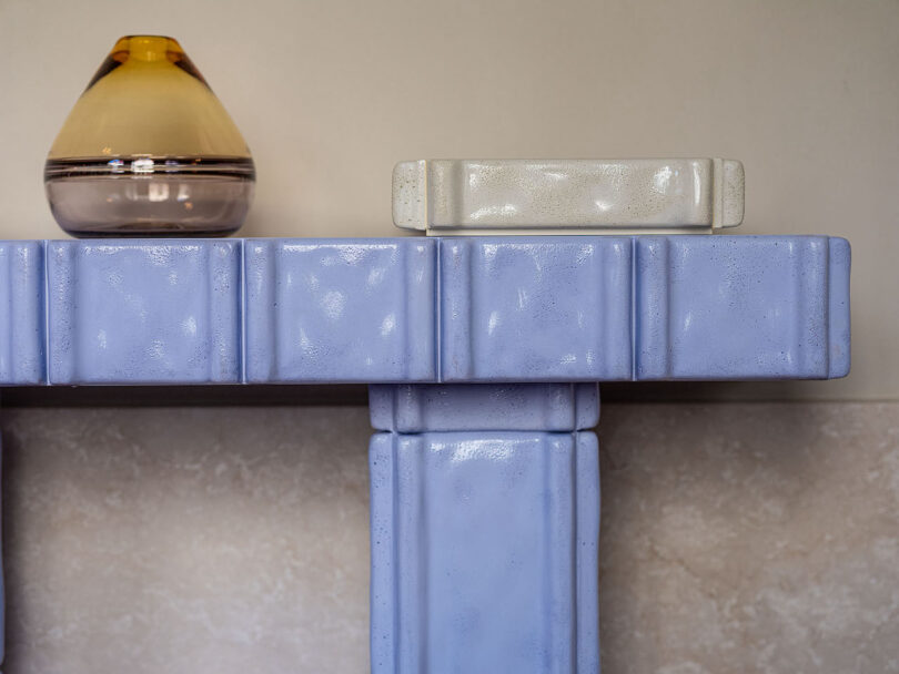 A close-up of a light purple, textured table with a brown-tinted glass vase and a rectangular white ceramic object placed on top
