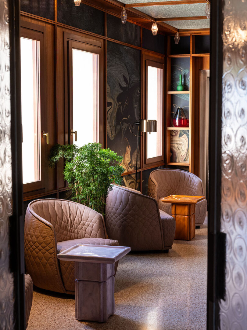 A modern lounge area with quilted armchairs, square tables, potted plants, and wood-paneled walls with large windows and built-in shelves