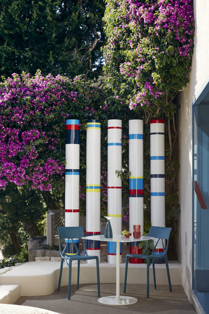 Outdoor patio with a round white table, two blue chairs, and four tall, white columns with colorful bands, surrounded by lush greenery and blooming purple flowers