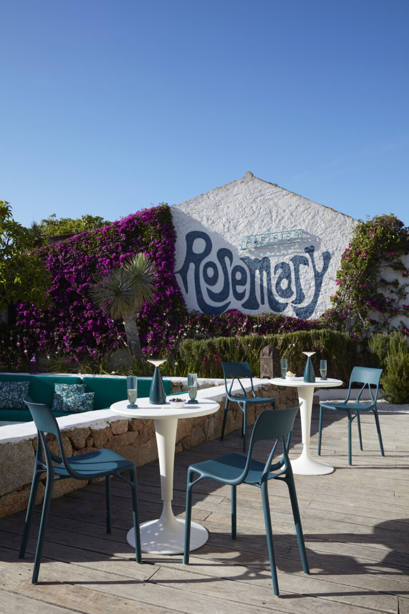 Outdoor patio with teal chairs and white tables set for dining; a white building with "Rosemary" painted on it and purple flowers on the wall in the background