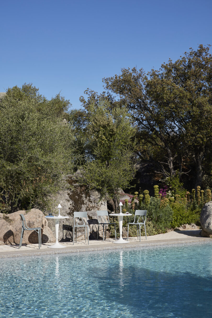 Poolside view with four metal chairs and two small white tables on a concrete patio, surrounded by greenery and trees under a clear blue sky