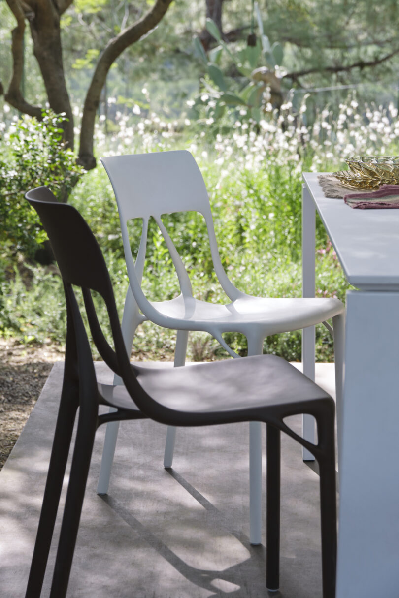 Two modern chairs, one black and one white, are placed next to a white outdoor table on a patio with greenery and trees in the background