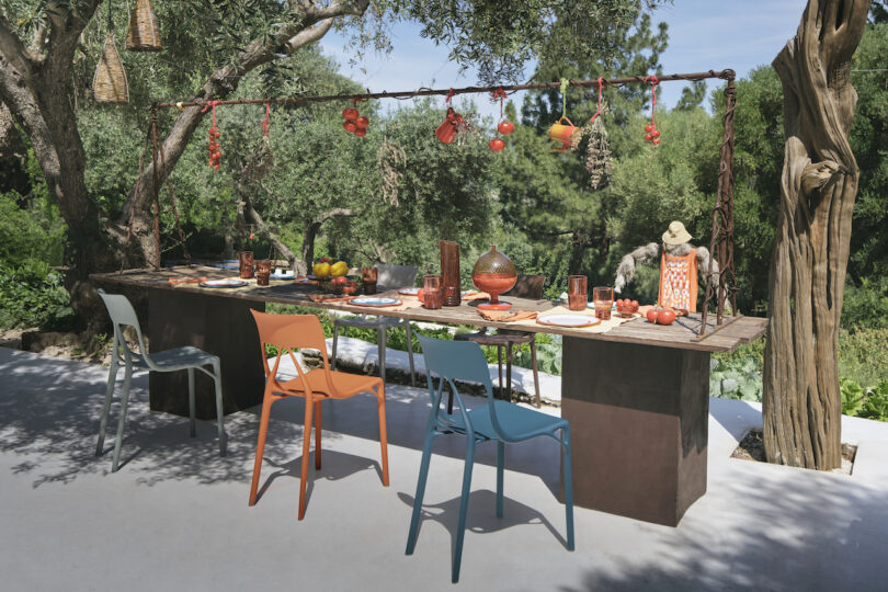 An outdoor dining table set for a meal, featuring colorful chairs, assorted tableware, hanging decorations, and surrounded by lush trees and greenery