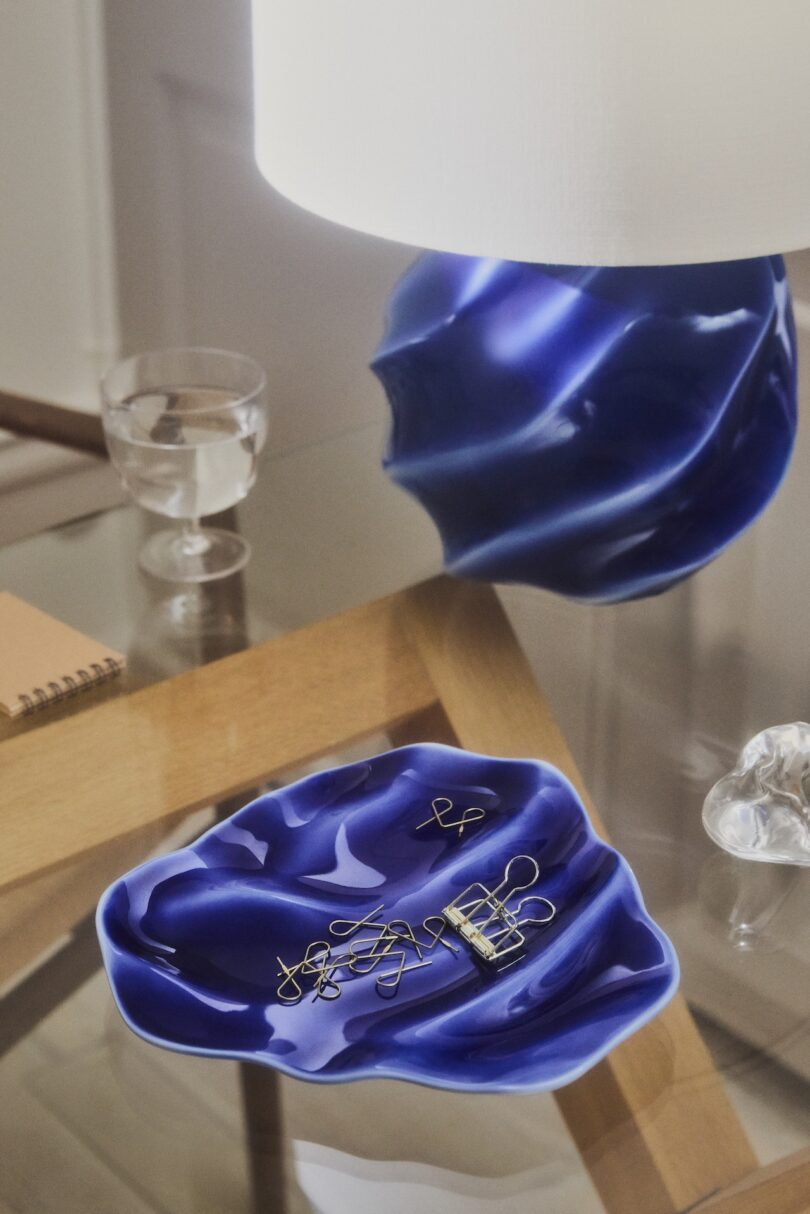 A blue wavy ceramic dish holding gold paper clips sits on a glass table beside a matching lamp, a glass of water, a notebook, and a clear decorative object